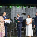 Na scenie stoi pięć osób. Kobieta trzyma chleb na białej serwecie. Tło ciemne, oświetlenie zielonymi reflektorami.