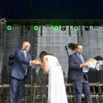 Grupa ludzi na scenie przy chlebach, elegancko ubrani, w tle czarny materiał i zielone światła sceny.