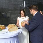 Na zdjęciu kobieta w białej sukni i mężczyzna w garniturze dzielą chleb. Na stole leży chleb ozdobiony ziołami i słonecznikami. Czarny materiał w tle.