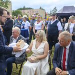 Na zdjęciu mężczyzna w garniturze oferuje chleb osobom siedzącym na krzesłach. Tło zielone, na zewnątrz, z namiotem i tłumem. Ludzie ubrani elegancko, atmosfera formalna.