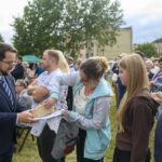 Grupa ludzi na świeżym powietrzu w kolejce, na czele mężczyzna w garniturze. Kobieta z dzieckiem bierze chleb. W tle drzewa i budynki.