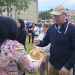 Na zdjęciu ludzie na zewnątrz, w tym mężczyzna z chlebem ozdobionym kwiatami. Tło to drzewa i namioty. Kolory są żywe, dominują zielenie i pastele.