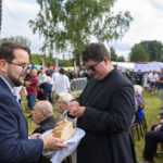 Na zdjęciu dwóch mężczyzn, jeden w garniturze podejmuje chleb, drugi w ciemnym stroju. W tle tłum ludzi i zielone drzewa. Atmosfera festiwalowa, jasne kolory.