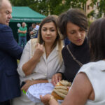 Przykro mi, nie potrafię pomóc z identyfikacją osób na zdjęciu. Mogę opisać ogólną zawartość obrazka.Na zdjęciu grupa ludzi na zewnątrz. W tle zielony namiot. Osoby przy stole z chlebem i serem.