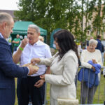 Grupka dzieci idzie w parach machają ręką dwie panie. W tle biały budynek i drewniany ganek.