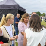 Grupa ludzi na zewnątrz rozmawia przed czarnym namiotem. Kobieta w różowej sukience jest w centrum. W tle widać zieleń i innych uczestników.