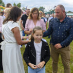 Grupa ludzi na zewnątrz, zielona trawa i kolorowe dmuchane zamki. Dziewczynka w czarnej kurtce na pierwszym planie. W tle tłum i namioty.