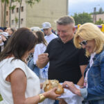 Na zdjęciu troje uśmiechniętych ludzi dzieli się chlebem. Tło to zielona trawa i kolorowe dmuchańce. Jest jasny dzień, ludzie są w casualowych strojach.
