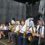 Orkiestra dęta na scenie gra na instrumentach, w tle szarość kurtyny. Muzycy w białych mundurach z czerwonymi kapeluszami. Dyrygent prowadzi zespół z lewej strony.