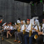 Orkiestra dęta na scenie, muzycy w białych koszulach i czerwonych czapkach. Dyrygent w granatowo-czerwonym mundurze. Czarne tło.