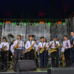 Na scenie stoi orkiestra dęta w białych koszulach i czapkach, z instrumentami. Tło jest ciemne, z kolorowymi światłami na górze. Po prawej stronie osoba wręcza dyplom.