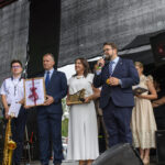 Grupa osób na scenie, w eleganckich ubraniach, trzyma nagrody. Tło to ciemny ekran. Obok stoi mężczyzna z mikrofonem.