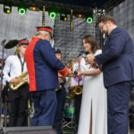 Na scenie stoi orkiestra dęta; osoby trzymają instrumenty. Kobieta w bieli i mężczyzna w garniturze podają coś starszemu mężczyźnie. Tło wypełnia ciemny materiał i kolorowe światła.