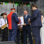 Na scenie mężczyzna w mundurze wręcza dyplom innemu mężczyźnie w garniturze. Obok stoją muzycy z saksofonami, ubrani na biało i czarno. Tło to ciemne zasłony i kolorowe światła sceniczne.