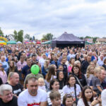 Na zdjęciu widać dużą grupę ludzi na festynie. W tle kolorowe dmuchańce i namioty. Niebo jest pochmurne.