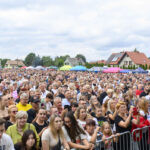 Tłum ludzi stoi na zewnątrz, w tle kolorowe stragany i domy. Pogoda jest pochmurna. Atmosfera festynu lub wydarzenia.