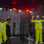 Dwóch mężczyzn w jaskrawozielonych strojach stoi na scenie. W tle ciemne zasłony i perkusja. Światła sceniczne świecą czerwonym i białym blaskiem.