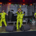 Trzech artystów w zielonych garniturach na scenie koncertowej. Za nimi czarne tło sceniczne i oświetlenie w kolorach. Na scenie widoczne są instrumenty muzyczne, w tym perkusja.