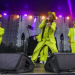 Trzech mężczyzn na scenie w jaskrawych zielonych garniturach. Na środku artysta z mikrofonem, tło kolorowe światła. Energetyczny nastrój koncertu.