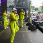 Trzech mężczyzn w neonowych zielonych strojach występuje na scenie. Tłum ludzi ogląda ich koncert na świeżym powietrzu. Tło jest ciemne, a w oddali widać drzewa i budynki.