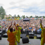 Koncert na świeżym powietrzu, czterech artystów w żółtych strojach, tłum ludzi, kolorowe namioty i drzewa w tle.