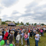 Duża grupa ludzi na trawniku podczas festiwalu na świeżym powietrzu. W tle kolorowe namioty i drzewa. Niebo jest pochmurne.