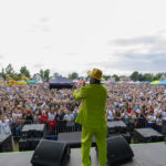 Mężczyzna w zielonym garniturze i żółtym kapeluszu występuje na scenie. Wielotysięczny tłum słucha pod chmurką. W tle dmuchane atrakcje i kolorowe namioty.