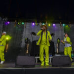 Na scenie trzech artystów w jasnozielonych strojach. Tło ciemne z kolorowymi światłami. Centralna postać śpiewa do mikrofonu.