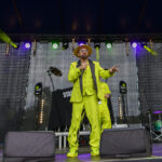 Na scenie wykonawca w jaskrawozielonym garniturze. Tło sceny jest ciemne, z kolorowymi światłami. Widoczne są instrumenty i sprzęt muzyczny.