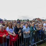Grupa ludzi stoi przy metalowych barierkach na otwartym powietrzu. Tło to domy i nieliczne drzewa pod pochmurnym niebem. Większość osób trzyma telefony w dłoniach.