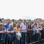 Na zdjęciu znajduje się tłum ludzi stojących za metalową barierką. Część osób robi zdjęcia telefonami, niebo jest jasne. W tle widać domy i namioty.