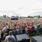 Na zdjęciu tłum ludzi na festiwalu pod gołym niebem. Tło to kolorowe namioty i drzewa. Przód sceny zabezpieczany przez ochroniarzy.