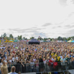 Tłum zgromadzony na festynie na otwartym polu. W tle widoczne kolorowe namioty i dmuchane atrakcje. Niebo częściowo zachmurzone.