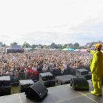 Scena koncertowa z artystą w żółtym stroju przed tłumem ludzi. Tło stanowi niebo i namioty festiwalowe. Atmosfera jest pełna energii i radości.