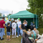 Stoisko Banku z zielonym namiotem na tle drzew i nieba. Tłum ludzi stoi, widać dzieci i dorosłych. Atmosfera piknikowa.