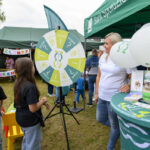 Na zdjęciu jest plenerowy festyn z zielonym namiotem. Kobieta obok koła fortuny z napisami. Balony i ulotki na stoliku w jasnych kolorach.