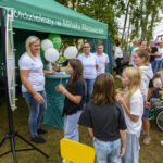 Grupa ludzi przed zielonym namiotem banku, ozdobionym balonami. Trawa na ziemi, drzewa w tle, dzień jest jasny. Dzieci patrzą na dorosłych.