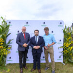 Trzej mężczyźni stoją przed białą ścianką z logo, na tle trawy. Ubrani elegancko, dominują kolory niebieski i zielony. Po bokach ścianki są dekoracje z roślin.