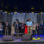 Na scenie stoi kilkanaście osób w formalnych strojach. Tło czarne, z efektami świetlnymi. Przemawia mężczyzna w błękitnej koszuli.