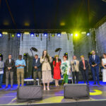 Grupa ludzi stoi na scenie pod niebieskim zadaszeniem, światła sceniczne świecą na żółto. Tło ciemne i teksturowane. Widać mikrofon i głośniki na scenie.