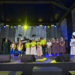 Na scenie grupa ludzi w kolorowych ubraniach. Tło ozdobione niebieskimi i żółtymi światłami. Jedna osoba trzyma znak 500 zł.