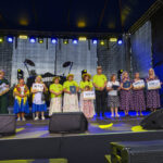 Na scenie stoi grupa ludzi w barwnych strojach, trzymających czeki. Tło stanowi czarny materiał, który kontrastuje z kolorowymi światłami. Po obu stronach sceny są prowadzący z mikrofonami.