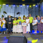 Grupa osób stoi na scenie, trzymając duże czeki. Kolory dominujące to zielony i niebieski. W tle widać ciemne oświetlenie sceny.