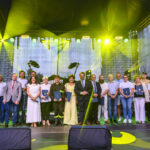 Zdjęcie przedstawia grupę ludzi na scenie w kolorowych ubraniach. W tle czarne, podświetlone kurtyny i żółte reflektory. Obecni trzymają dyplomy, co sugeruje ceremonię wręczenia nagród.