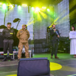 Na scenie czterech ludzi, z tyłu czarny ekran z napisem. Światła w tonacji żółtej i zielonej. Jeden z mężczyzn przemawia.