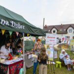 Na zdjęciu widać stoisko z zielonym namiotem z napisem Gmina Dobre. Kolorowe girlandy wiszą w tle, a na wystawie są obrazy i dekoracje. Ludzie rozmawiają i oglądają sztukę, obok stoi biały dom.