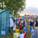 Ludzie na pikniku w otoczeniu zielonych namiotów. W tle widać atrakcje na placu zabaw. Wieczorne niebo jest lekko pochmurne.