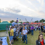 Tłum ludzi na festynie na świeżym powietrzu. Namioty w kolorach zielonym i pasiastym. W tle zjeżdżalnie i kolorowe balony.