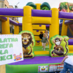 Dzieci bawią się na kolorowym dmuchanym zamku z żółtymi i fioletowymi elementami. Tło przedstawia kolorowe zjeżdżalnie z motywem zoo. Na pierwszym planie jest tabliczka BEZPŁATNA STREFA DLA DZIE