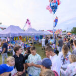 Na zdjęciu widać kolorowy jarmark z tłumem ludzi. Stragany mają niebiesko-białe daszki, w tle widać różową karuzelę. Balony helowe unoszą się nad głowami uczestników.